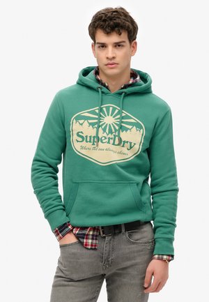 Grüne Kapuzensweatshirt mit einem großen cremefarbenen Grafik, die Berg- und Sonnendetails zeigt. Getragen über einem karierten Hemd, kombiniert mit grauen Jeans.