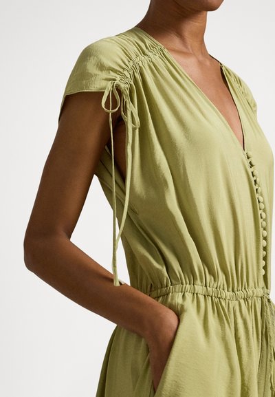 Γυναίκα με αμάνικο φόρεμα σε ανοιχτό ελιάς χρώμα, με υφασμάτινα κουμπιά μπροστά, ελαστική μέση με πτυχώσεις και κορδόνια �στους ώμους, με το χέρι στην τσέπη.