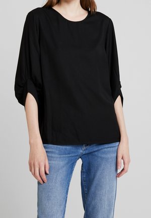 Mujer con una camiseta holgada negra de manga tres cuartos y vaqueros azules sobre un fondo gris.