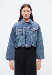 Közép kék farmerdzseki klasszikus gallérral, elöl gombos záródással, két mellzsebbel és túlméretezett, volumentus ujjakkal.