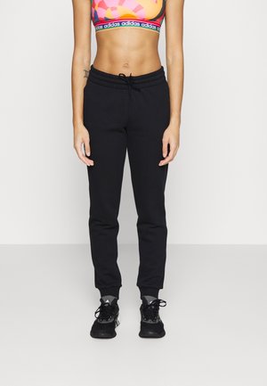 Athletische Person trägt bunten Sport-BH, schwarze Jogginghosen mit Kordelzug und schwarze Adidas-Sneaker und steht vor einem schlichten Hintergrund.