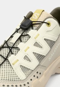 Ελαφρύ αθλητικό παπούτσι με άνω μέρος από δίχτυ σε κρεμ χρώμα, μπεζ λεπτομέρειες και λαστιχένια σόλα. Διαθέτει ελαστικά κορδόνια και λογότυπο στο πλάι.