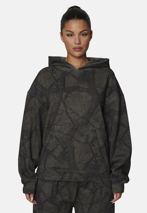 Vrouw met donker haar in een staart draagt een losse donkergrijze hoodie en bijpassende broek met een subtiel abstract takkenpatroon.
