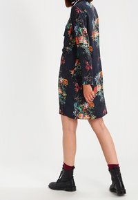 Kwiatowa sukienka z ciemnej tkaniny z żywymi nadrukami botanicznymi. Długie rękawy, kołnierz w paski, w komplecie z czarnymi sznurowanymi botkami.