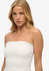 Biała sukienka bez ramiączek z marszczonym topem i falbanką przy dekolcie, wykonana z teksturowanej tkaniny o dopasowanej sylwetce.