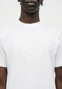 Άτομο που φορά λευκό μπλουζάκι με ανυψωμένο, λευκό-σε-λευκό, τριγωνικό λογότυπο Guess και κείμενο στο στήθος, σε απλό φόντο.