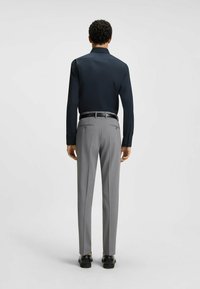 Marinblå skjorta med knappar och långa ärmar, matchad med ljusgrå mönstrade byxor och svarta skor, sedd bakifrån.