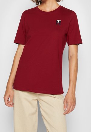 Persoon draagt een effen donkerrood T-shirt met korte mouwen en een klein "TH"-logo op de borst, gecombineerd met lichtbeige broek.