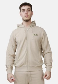 Felpa con zip color beige, dotata di cappuccio e realizzata in tessuto morbido; presenta un piccolo logo verde sul petto e polsini e orlo a coste.
