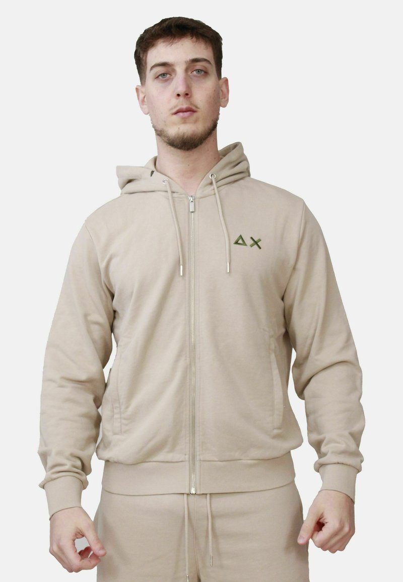 Felpa con zip color beige, dotata di cappuccio e realizzata in tessuto morbido; presenta un piccolo logo verde sul petto e polsini e orlo a coste.