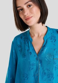 Camisa de botones en satén teal con estampado floral, mangas cortas, cuello de muesca, bolsillo en el pecho y botones en contraste. Textura suave.