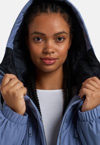 Veste isolante bleue avec capuche, dotée d'une doublure intérieure douce et de poignets élastiques. Le modèle tient la veste ouverte pour la présentation.