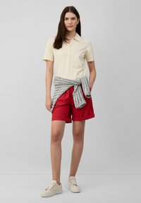 Polo shirt jaune clair avec un col, shorts rouges, pull en maille gris noué à la taille, et baskets beiges. Tenue simple et décontractée.