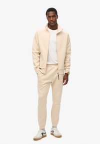 Chaqueta beige de cremallera sobre una camiseta blanca, combinada con pantalones tapered beige a juego. Zapatillas con base blanca y acentos negros.