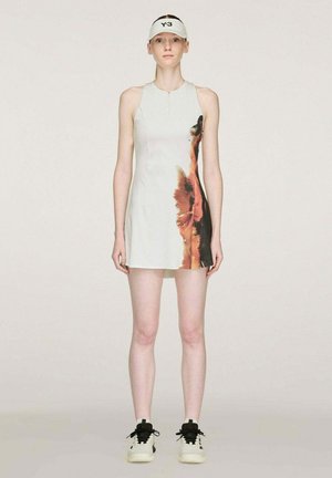 Mujer con vestido blanco sin mangas con gráfico marrón en el lado derecho, visera blanca y zapatillas blancas y negras, de pie contra un fondo liso.