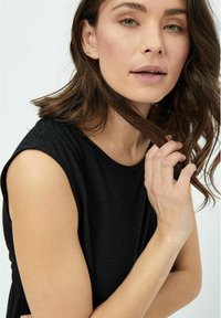 Čierny top bez rukávov zo mäkkej tkaniny, s okrúhlym výstrihom a jemným drapériom. Modelka si ľahko drží vlasy, predvádzajúc prirodzený make-up a svetlý tón pleti.