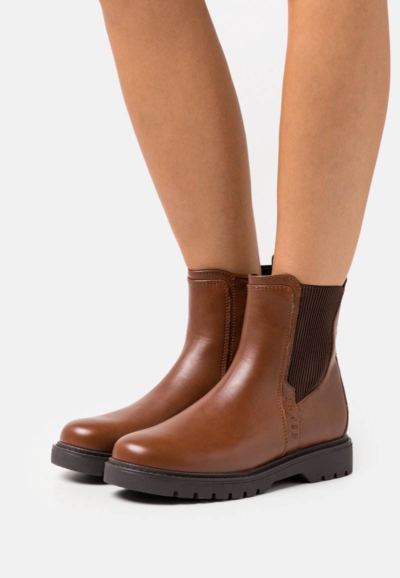 Bottines en cuir marron avec panneaux latéraux élastiques noirs, portées sur jambes nues, avec semelles épaisses en caoutchouc noir et bouts arrondis.