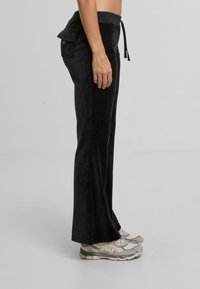 Dopasowane czarne spodnie z weluru z elastycznym paskiem w talii i boczną kieszenią, z fasonem rozszerzających się nóg i miękką fakturą. Noszone z jasnymi sneakersami.