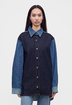 Femme aux longs cheveux foncés portant une veste en jean bicolore avec des manches et un col bleu clair, debout devant un fond uni.