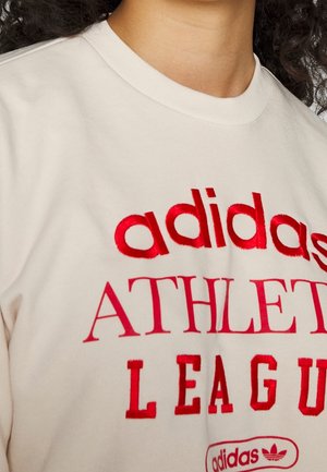 Zbliżenie osoby noszącej kremową koszulę z czerwonym haftowanym napisem „adidas ATHLETIC LEAGUE” z przodu.
