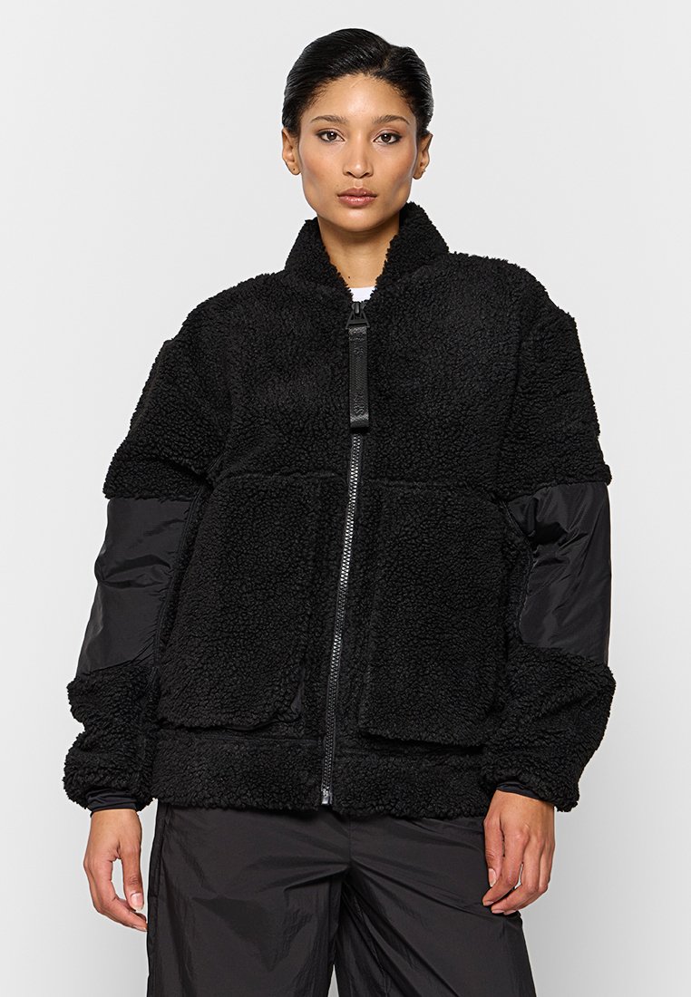 Veste en polaire noire avec des manches longues matelassées, fermeture éclair à l'avant et deux grandes poches à patch. Caractérisée par un extérieur texturé et doux.