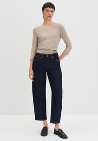 Maglietta beige a maniche lunghe con scollatura ampia, abbinata a jeans dritti blu scuro. Il modello indossa mocassini neri. Design semplice e minimal.