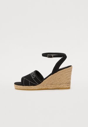 Sandales compensées noires avec une tige en toile, bride de cheville ajustable et talon compensé en jute. Semelle noire plate avec des détails minimalistes.
