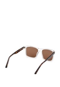 Silmät läpi näkyvät aurinkolasit, joissa on ruskeat linssit ja kilpikonnakuosiset varret. Kulmikas muotoilu, joka on tukeva ja kiillotteinen.