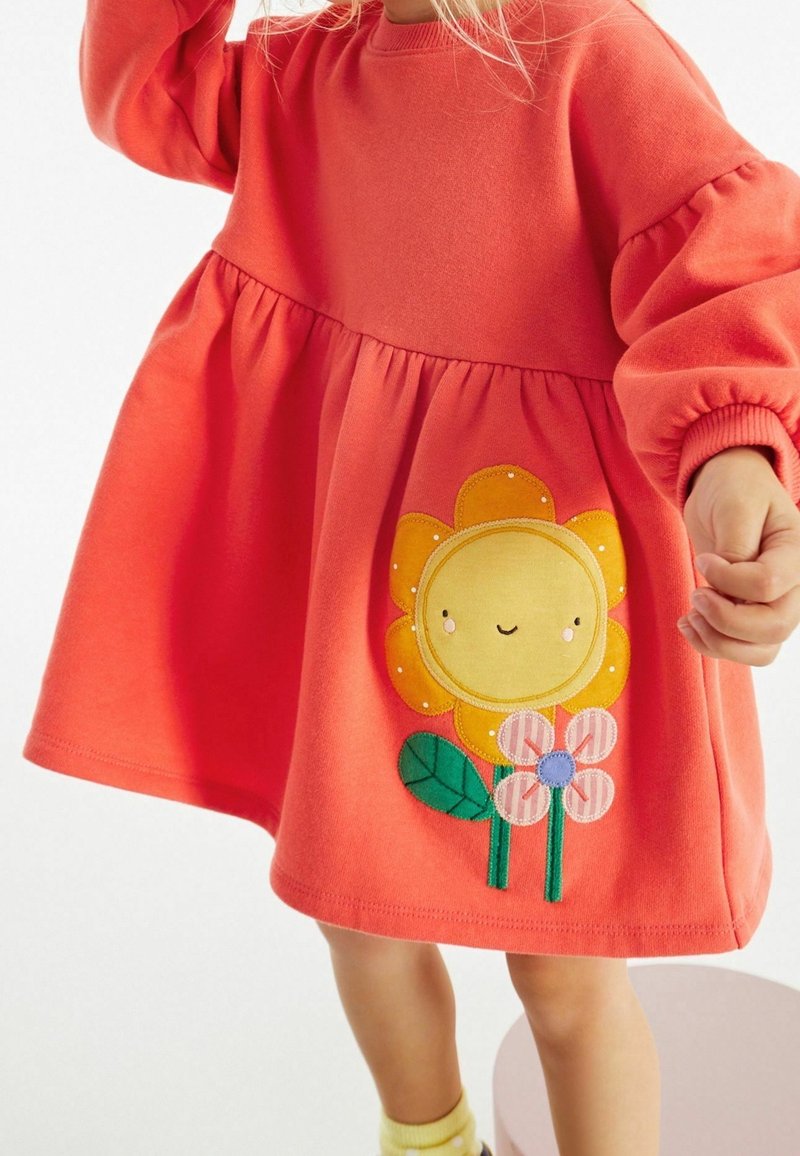 Enfant portant une robe corail avec de longues manches bouffantes et une fleur jaune souriante avec deux petites fleurs brodées sur la jupe.
