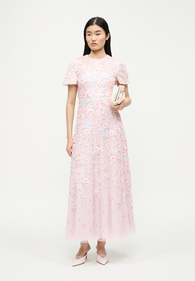 Γυναίκα με ροζ φόρεμα με κεντήματα από λουλούδια μέχρι το δάπεδο κρατάει ένα λευκό τσαντάκι, φοράει ροζ μυτερές γόβες σε λευκό φόντο.