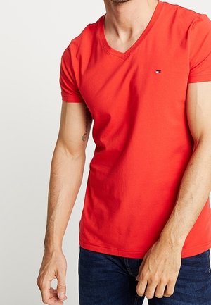 Hombre con camiseta roja de manga corta con cuello en V y pequeño logo en el pecho, y vaqueros azul oscuro sobre un fondo liso.
