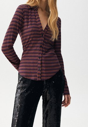 Woman wearing a patterned purple and orange buttoned shirt with black sequined pants, standing against a plain white background.