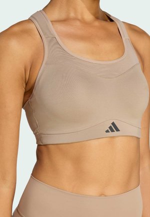Beige sportbeha met brede bandjes, een gladde textuur, een gebogen halslijn en zwart logo-detail aan de onderkant. Geschikt voor sportieve doeleinden.