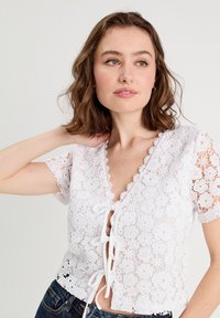 Haut en dentelle blanche avec motifs floraux, manches courtes et fermeture à nouer à l'avant. Design court avec un encolure et un ourlet festonnés.