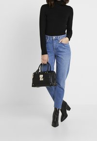 Zwarte leren handtas met een gestructureerd oppervlak en gouden hardware, vastgehouden in een hand; gecombineerd met een zwarte coltrui en blauwe jeans.