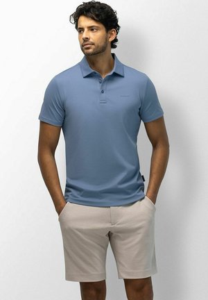Man met krullend haar die een blauwe polo met korte mouwen en beige korte broek draagt, staat met handen in de zakken en kijkt opzij.