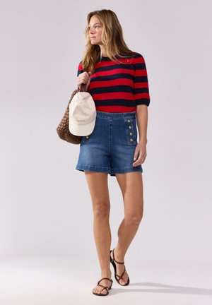 Maglione a righe rosse e blu, pantaloncini di jeans blu con bottoni laterali, borsa in pelle intrecciata marrone e un cappellino beige chiaro. Abbigliamento estivo casual.