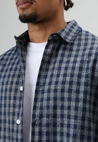 Marineblaue und graue kariertes Hemd mit Knopfkragen, weißen Knöpfen und weicher gewebter Struktur über einem weißen Unterhemd.