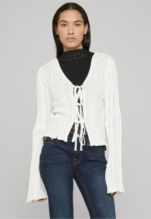 Weißer Strickcardigan mit tiefem V-Ausschnitt, Kordelzugverschluss und gerippter Textur. Lange Ärmel werden an den Handgelenken schmaler, getragen über einem schwarzen Oberteil.