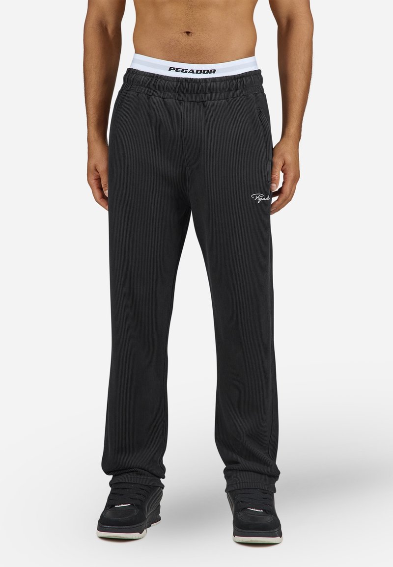 Pantalon de survêtement noir côtelé avec une ceinture élastique, arborant un logo blanc PEGAADOR. Le tissu est lisse et la coupe est décontractée.