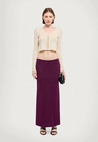 Kvinne iført en kremfarget kort cardigan, lilla høytlivs maxi skjørt, svarte sandaler, og holder en liten svart clutch.