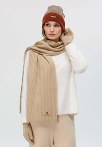 Sciarpa lavorata a maglia beige, berretto a coste marrone, guanti beige aderenti e maglione bianco morbido; tutti realizzati con materiali soffici e con una finitura testurizzata.