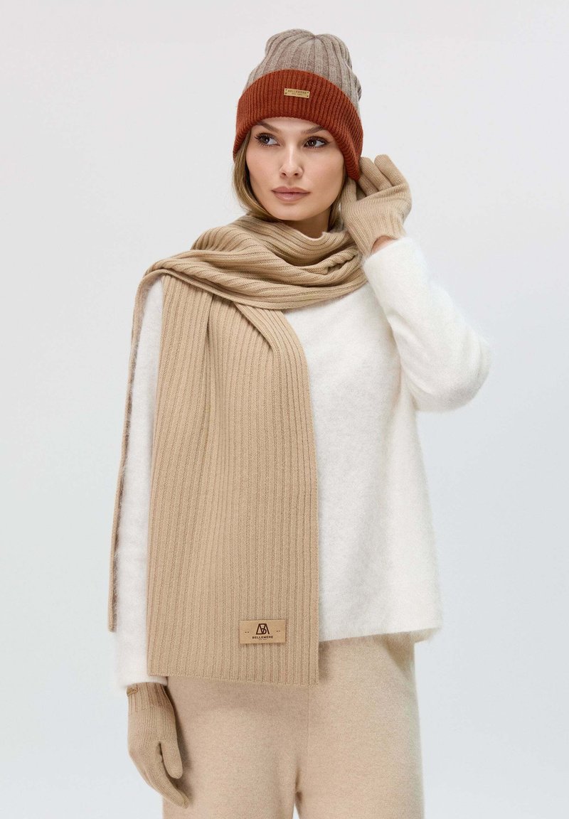 Sciarpa lavorata a maglia beige, berretto a coste marrone, guanti beige aderenti e maglione bianco morbido; tutti realizzati con materiali soffici e con una finitura testurizzata.