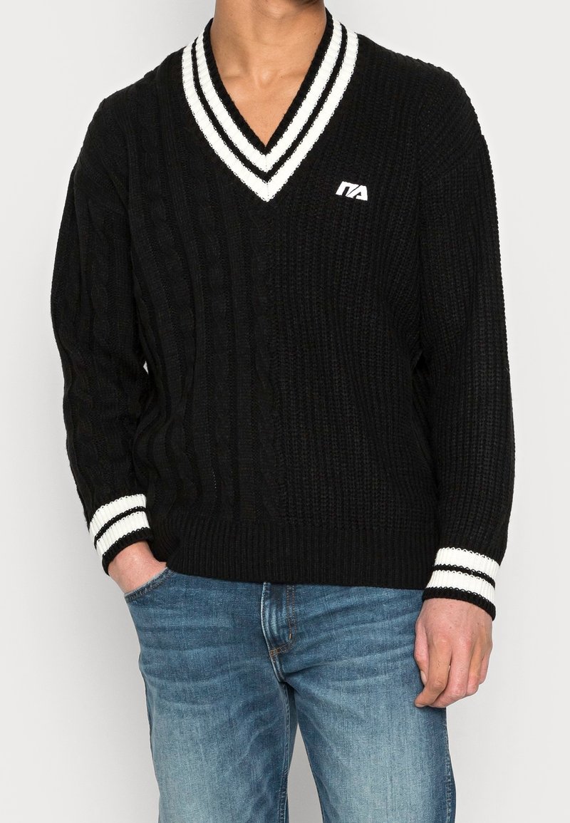 Uomo che indossa un maglione a V in maglia cablè nera con strisce bianche sul colletto e sui polsini, abbinato a jeans di denim blu.