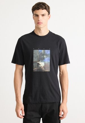 Junger Mann trägt ein schwarzes T-Shirt mit einem bunten Grafik eines sitzenden Menschen in einem dunklen, atmosphärischen Raum, darüber das Wort "HUGO".
