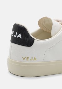 Hvite lærersneakers med en svart hæltab som har hvit tekst "VEJA" og en gull logo. Glatt tekstur med et minimalistisk design.