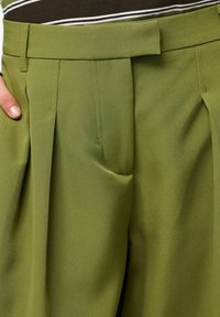 Vihreät räätälöidyt housut, joissa on laskokset, sileä tekstuuri ja vyötärönauha. Suunnitteluun integroidut taskut parantavat toiminnallisuutta.