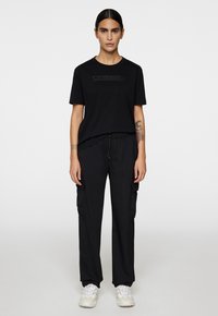 Czarna koszulka z teksturowanym logo na piersi, zestawiona z czarnymi spodniami cargo z bocznymi kieszeniami i elastycznym pasem na sznurku. Białe sneakersy.