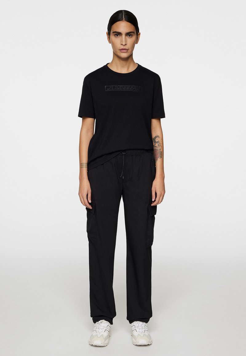 T-shirt nera con logo testurizzato sul petto, abbinata a pantaloni cargo neri con tasche laterali e vita con coulisse. Sneakers bianche.