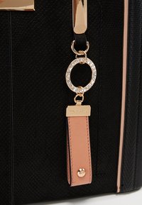 Musta teksturoidulla käsilaukulla, jossa on kultaiset yksityiskohdat. Laukussa on kristallikoristeltu pyöreä charm ja vaaleanpunainen nahkatagi logolla.